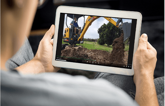 Hands holding a tablet displaying a Yuchai Americas excavator digging soil on a construction site